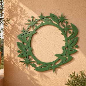 Könnte beinhalten: Ein grüner Metallkranz mit kreisförmigem Design, mit stilisierten Blättern, Blumen und sternförmigen Formen. Der Kranz ist an einer strukturierten, hellbraunen Wand montiert, mit Schatten von Pflanzen in der Nähe.