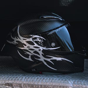 Peut inclure: Un casque de moto en fibre de carbone noir avec un motif graphique tribal argenté sur le côté. Le casque comprend une visière teintée foncée avec un fin motif de fil de fer barbelé argenté imprimé sur le bord supérieur de la visière.