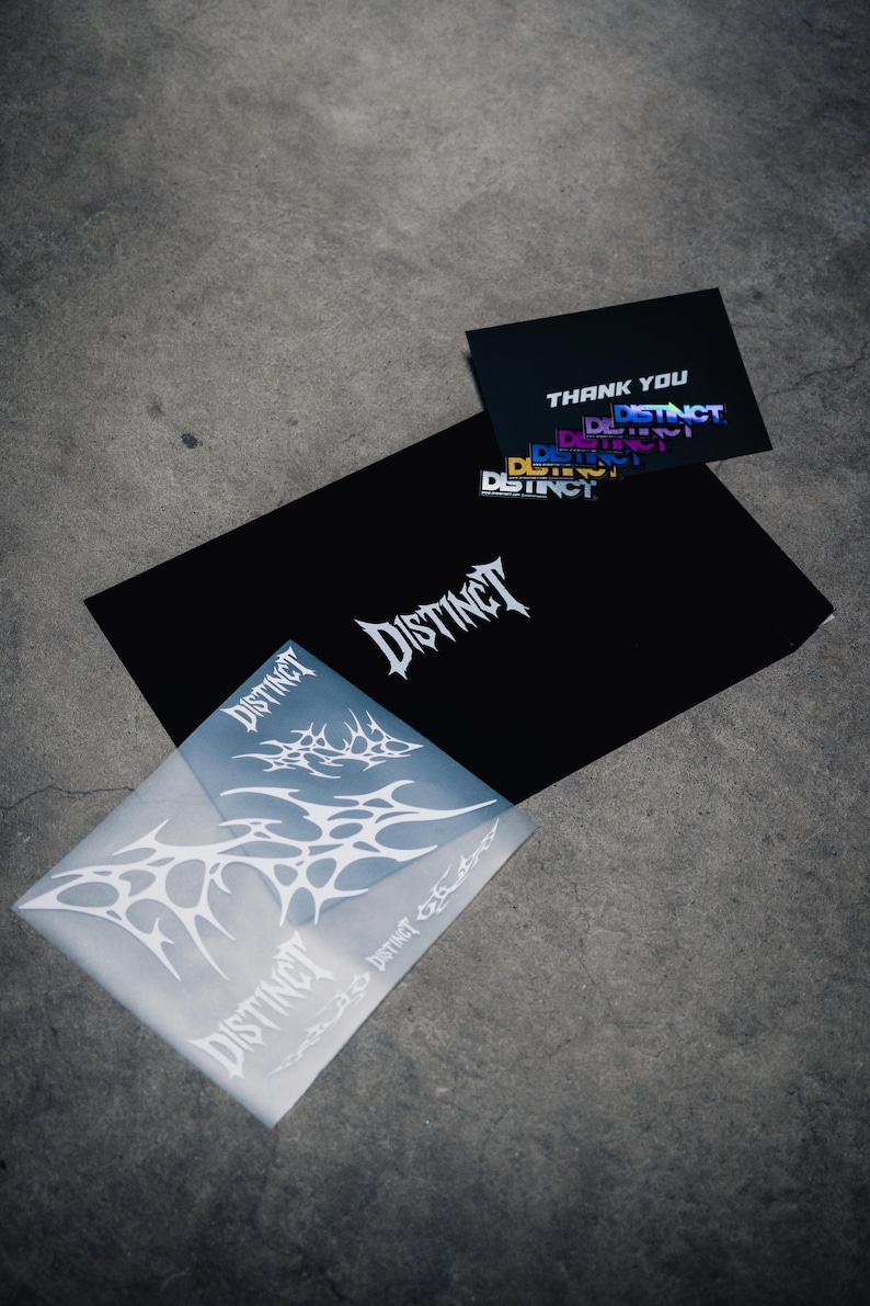 May include: A collection of stationery items featuring the word "DISTINCT" in a stylized font. The items include a black envelope, a translucent envelope, and a thank you card, all with the same branding. The background is a gray concrete surface.