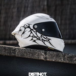 May include: A white motorcycle helmet with a black visor and tribal-style black graphics. The helmet is resting on a concrete surface. The text "DISTINCT. DESIGNED TO BE SEEN" is visible at the bottom.