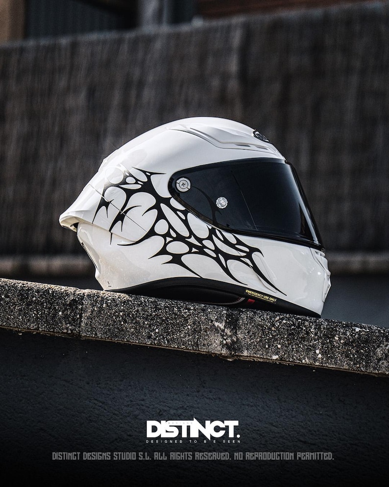 May include: A white motorcycle helmet with a black visor and tribal-style black graphics. The helmet is resting on a concrete surface. The text "DISTINCT. DESIGNED TO BE SEEN" is visible at the bottom.