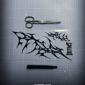 May include: A top-down shot shows a gray cutting mat with a pair of scissors, a black utility knife, and black tribal-style decals. The decals are in a spiky, abstract design. The word "DISTINCT" is printed on the bottom.