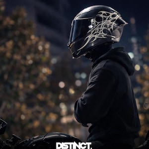 May include: A black motorcycle helmet with a decorative white design. The helmet has a tinted visor and is worn by a person in a black hoodie. The image includes the text "DISTINCT. DESIGNED TO BE SEEN."