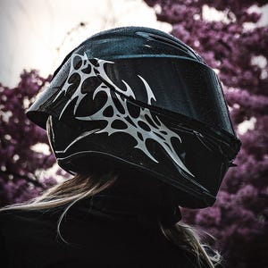 May include: A black motorcycle helmet with a silver tribal design. The helmet has a dark visor and is wet with raindrops. The background features blurred purple flowers.