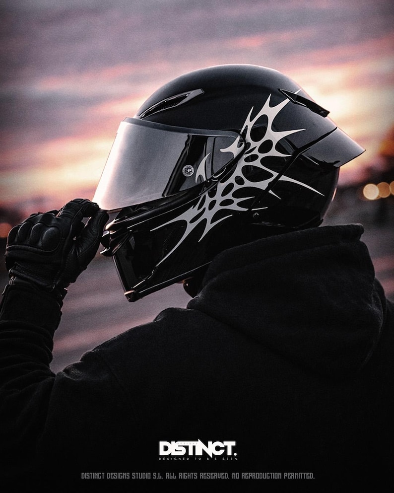 May include: A glossy black motorcycle helmet with a silver tribal design on the side. The helmet has a tinted visor and is held by a gloved hand. The word "DISTINCT" is printed at the bottom.
