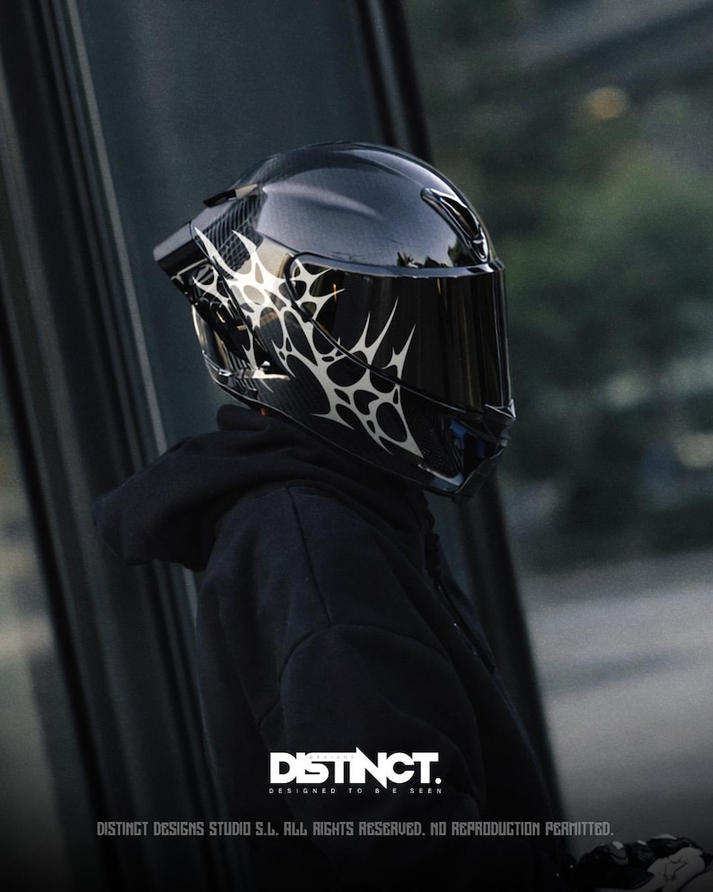 May include: A glossy black motorcycle helmet with a dark visor and a white, abstract design on the side. The helmet is part of a motorcycle gear collection. The text "DISTINCT. DESIGNED TO BE SEEN" is visible at the bottom.