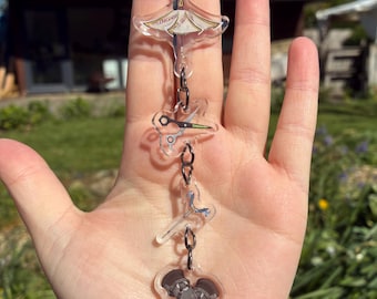 May include: A clear acrylic keychain with multiple charms. The charms include a kite, scissors, a pair of scissors, and a heart-shaped charm with a gray illustration. The keychain is attached to a silver chain.