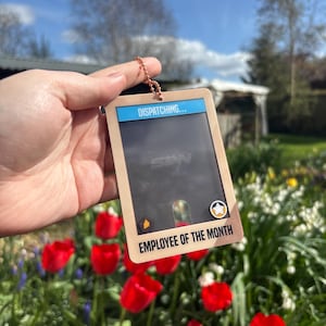 May include: A rectangular employee badge with the text "DISPATCHING..." at the top and "EMPLOYEE OF THE MONTH" at the bottom. The badge is held up in front of a background of red tulips and greenery.