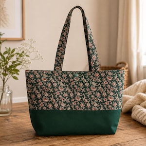 May include: A tote bag featuring a dark green base and a top section patterned with a dense floral and paisley print in shades of red, white, and green. The bag includes two matching fabric shoulder straps and a single green snap button closure.