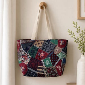 May include: A quilted tote bag featuring a patchwork design with various floral, paisley, and geometric patterns in shades of deep red, navy blue, and forest green. The bag has two off-white fabric handles and a rectangular, structured shape.