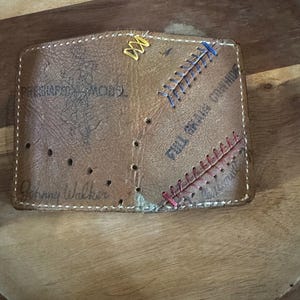 May include: A brown leather wallet with stitched accents in yellow, blue, and red thread. The wallet has the words "Johnny Walker" and "PRESHAPED MACHO" printed on it. The wallet is on a wooden surface.
