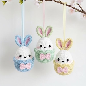 Può includere: Tre ornamenti a forma di uovo di Pasqua all'uncinetto fatti a mano con orecchie da coniglio, ciascuno in un colore pastello diverso: blu, verde e giallo. Ogni uovo ha una faccia sorridente e un fiocco. Sono appesi a nastri.