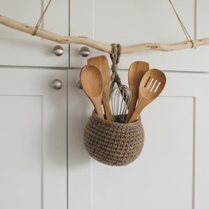 May include: A hanging kitchen utensil holder made of a woven, brown material. The holder is suspended from a natural wood branch and contains wooden spoons and a whisk. The background features white cabinets.