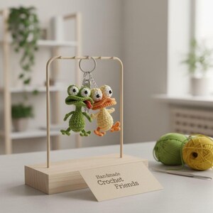 Puede incluir: Dos llaveros de animales hechos a mano con ganchillo: una rana verde con una lengua roja y un pato amarillo con detalles en naranja. Cuelgan de un marco de metal sobre una base de madera. Un letrero dice "Handmade Crochet Friends."