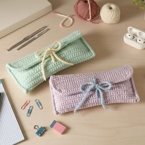 May include: Two handmade crochet cases in pastel colors. One is light green with a cream-colored bow, and the other is lavender with a light blue bow. The cases are rectangular with a flap closure. Crochet hooks, yarn, and office supplies are also visible.