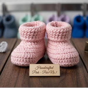 May include: A pair of handcrafted pink crochet baby booties. The booties are made of a soft, textured yarn and have a rolled cuff. A wooden sign in front of the booties reads "Handcrafted Pink - Pair No.1". Other baby booties are visible in the background.