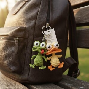 Puede incluir: Dos llaveros de ganchillo hechos a mano: una rana verde con lengua roja y un pato amarillo con pico y patas naranjas. Los llaveros est&aacute;n sujetos a una mochila de cuero marr&oacute;n. Una peque&ntilde;a etiqueta dice "Handmade Charm".