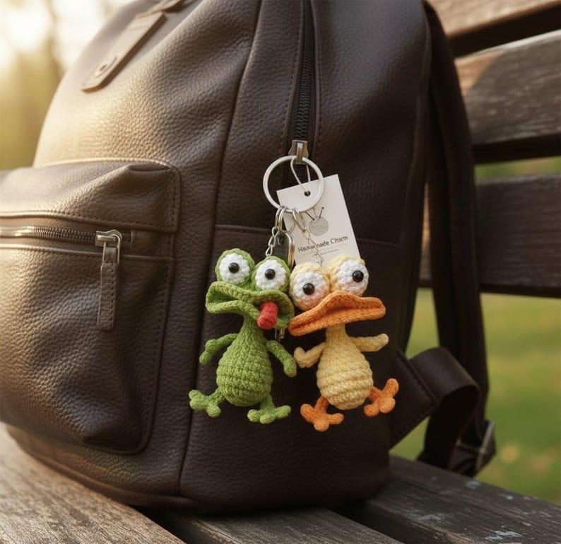 Puede incluir: Dos llaveros de ganchillo hechos a mano: una rana verde con lengua roja y un pato amarillo con pico y patas naranjas. Los llaveros est&aacute;n sujetos a una mochila de cuero marr&oacute;n. Una peque&ntilde;a etiqueta dice "Handmade Charm".