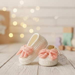 May include: A pair of handmade, white crochet baby shoes with peach-colored bows. The shoes are displayed on a light wooden surface. A small tag reads "Handmade with Lisa".