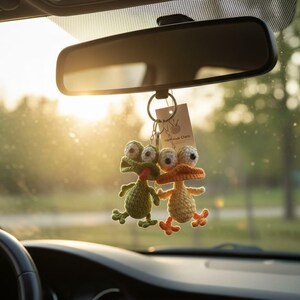 Puede incluir: Dos amuletos de coche hechos a mano, de ganchillo, cuelgan de un espejo retrovisor. Uno es una rana verde, el otro es un pato naranja. Ambos tienen ojos grandes y saltones y est&aacute;n unidos a un llavero con una peque&ntilde;a etiqueta que dice "Handmade Charm".