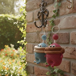 May include: Two handmade crochet keychains. One is teal with a tan brim and the other is red with a brown brim. Both have a round head and a small ball on top. They hang from a decorative hook on a brick wall.