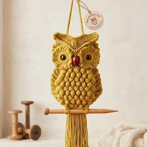 Op de afbeelding: Een handgemaakte, mosterdgele gehaakte uil voor aan de muur. De uil heeft grote zwarte ogen, een rode snavel en zit op een houten deuvel. Een houten label leest "Boho Wisdom Owl, Handmade with Love."