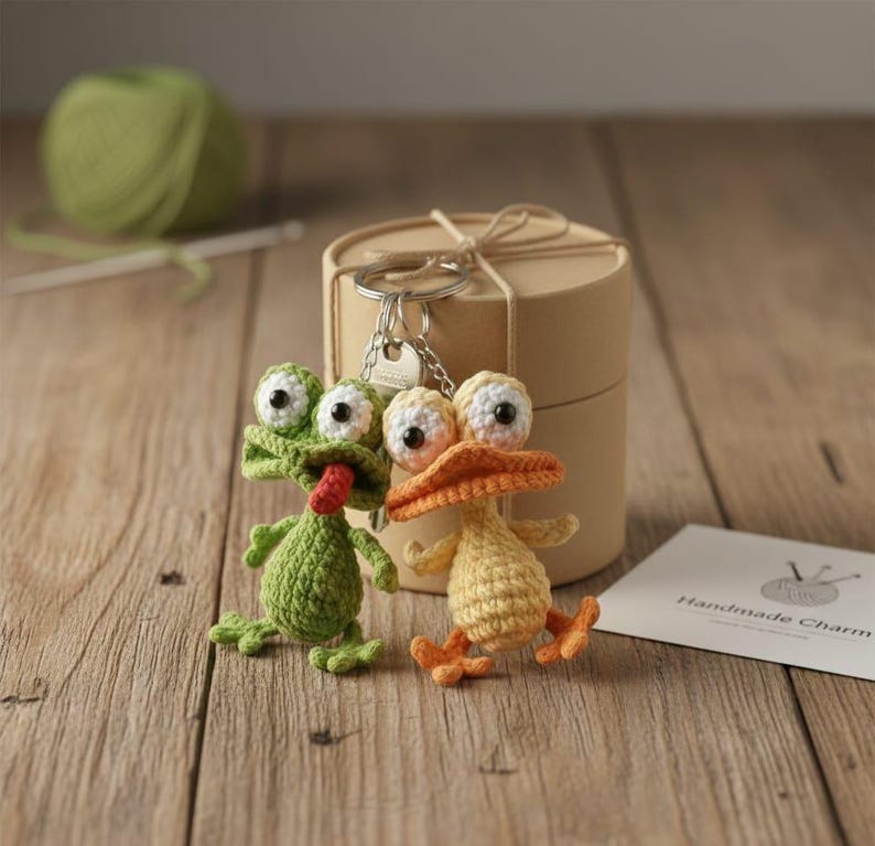 Puede incluir: Dos llaveros de ganchillo hechos a mano: una rana verde con una lengua roja y un pato amarillo con pico y patas naranjas. Los llaveros est&aacute;n unidos a un llavero plateado, junto a una peque&ntilde;a caja de regalo y una tarjeta que dice "Handmade Charm".