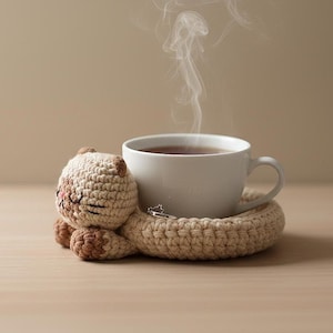 May include: A white ceramic teacup filled with tea, resting on a beige crocheted cat-shaped coaster. The cat has brown accents and closed eyes. Steam rises from the cup, suggesting the tea is hot. A small silver ring rests on the coaster.