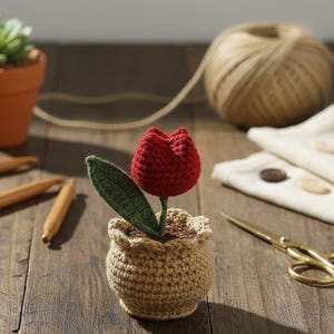 May include: A handmade crochet tulip in a small pot. The red tulip has a green leaf and sits in a beige pot. The pot is filled with brown material, and the background includes a ball of twine and a pair of gold scissors.