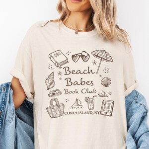 Peut inclure: T-shirt ivoire avec un motif marron représentant "Beach Babes Book Club" et des illustrations sur le thème de la plage. Le t-shirt est de coupe ample et est porté avec une veste en jean bleu clair.