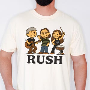Peut inclure: Un t-shirt à manches courtes de couleur crème avec une illustration de dessin animé représentant un groupe de trois musiciens jouant de leurs instruments. Sous les personnages, le mot RUSH est imprimé en grandes lettres capitales noires et grasses.