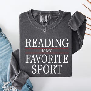Op de afbeelding: Een antracietgrijs sweatshirt met lange mouwen en ronde hals, voorzien van de witte tekst "READING IS MY FAVORITE SPORT" met dunne rode horizontale lijnen boven en onder het woord "IS MY". Het kledingstuk heeft een geribbelde halslijn.