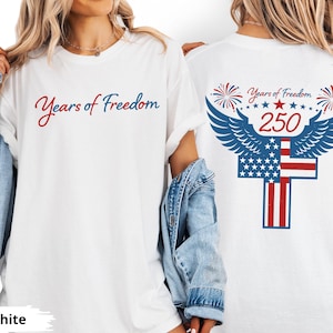 Peut inclure: T-shirt blanc avec l'inscription "Years of Freedom" en script rouge et bleu. Le dos présente un motif patriotique avec des ailes, des étoiles, des feux d'artifice et le chiffre "250" en rouge, ainsi qu'un drapeau américain.