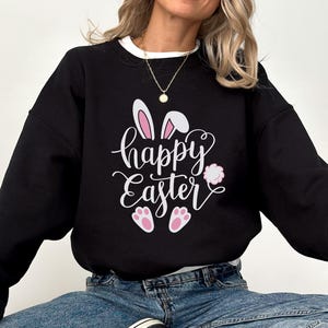 Peut inclure: Sweat-shirt noir avec l'inscription "happy Easter" en blanc, orné d'oreilles de lapin, de pattes et d'une queue touffue roses. Le sweat-shirt a un col blanc et est porté avec un jean bleu et des baskets noires.