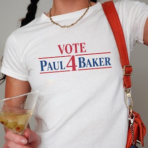 Könnte beinhalten: Ein weißes kurzärmeliges T-Shirt mit dem in Rot und Blau aufgedruckten Text VOTE PAUL 4 BAKER auf der Brust. Das T-Shirt wird mit einer leuchtend orangefarbenen Umhängetasche mit goldfarbenem Metallriemen und einem kleinen würfelförmigen Anhänger kombiniert.