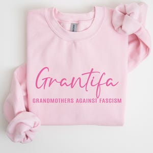 Peut inclure: Un sweat-shirt à col rond rose clair avec le mot Grantifa écrit en lettres cursives rose foncé, accompagné du texte GRANDMOTHERS AGAINST FASCISM imprimé en lettres bâton rose foncé juste en dessous sur la poitrine du vêtement.