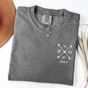 Peut inclure: Un t-shirt gris Comfort Colors avec un motif graphique blanc représentant des flèches, les lettres "H" et "O", les chiffres "31", "07" et "2014". Un collier argenté avec un petit pendentif est sur le t-shirt. Un chapeau marron est en arrière-plan.