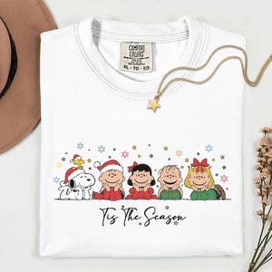 Peut inclure: Un t-shirt blanc avec une illustration des personnages Peanuts portant des tenues de fête, notamment des bonnets de Noël et des nœuds. Le texte 'Tis The Season' est imprimé sous les personnages, entouré de petites étoiles et flocons.