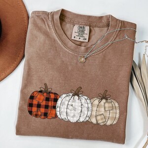 Peut inclure: Un t-shirt marron à manches courtes avec un motif de trois citrouilles aux imprimés variés : un à carreaux rouges et noirs, un à motifs floraux blancs et gris, et un à carreaux beiges. Le t-shirt porte une étiquette de la marque Comfort Colors.