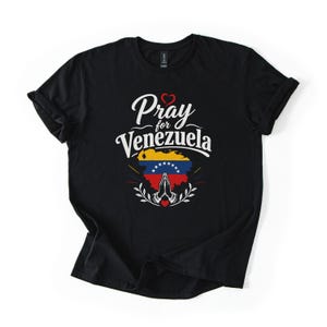 May include: A black short-sleeved t-shirt featuring the white text Pray for Venezuela above a graphic of the Venezuelan flag shaped like a map. Below the map are white praying hands, a small red heart, and decorative white leaf-like accents.