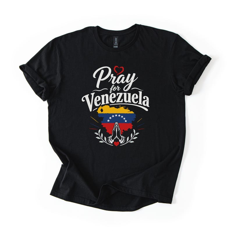 May include: A black short-sleeved t-shirt featuring the white text Pray for Venezuela above a graphic of the Venezuelan flag shaped like a map. Below the map are white praying hands, a small red heart, and decorative white leaf-like accents.