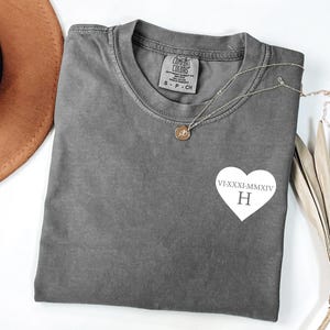 Peut inclure: Un t-shirt gris avec un motif blanc en forme de cœur avec des chiffres romains et la lettre "H". Un collier doré avec un pendentif repose sur le t-shirt. Le t-shirt est plié sur une surface blanche.