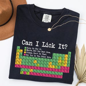 Peut inclure: Un t-shirt noir avec un graphique blanc représentant un tableau périodique des éléments et le texte "Can I Lick It?" au-dessus. Le graphique comprend une légende codée par couleur avec les phrases "Sure, Go For It," "Maybe, Not the Best Idea," "Please, Don't Do That," et "See You In The Other Side."