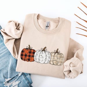 Peut inclure: Un sweat-shirt beige à col rond orné d'un motif représentant trois citrouilles alignées. La première citrouille présente un motif à carreaux rouges et noirs, la deuxième est blanche avec un imprimé de feuilles grises, et la troisième est beige.