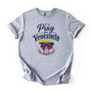 May include: A heather gray short-sleeve t-shirt featuring the dark blue text Pray for Venezuela above a map of Venezuela filled with the national flag colors of yellow, blue, and red, with a pair of praying hands and small red hearts.