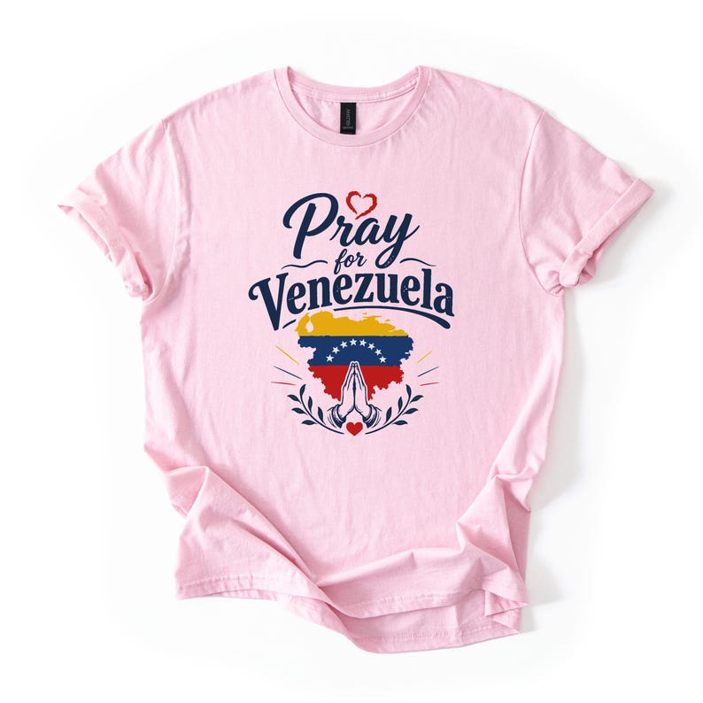 May include: A light pink t-shirt featuring a graphic print with the text Pray for Venezuela in dark blue script and block lettering. Below the text is a map of Venezuela in yellow, blue, and red with white stars, and a pair of praying hands.