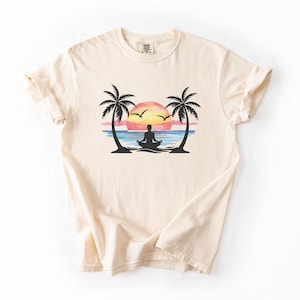 Peut inclure: T-shirt crème avec un motif de coucher de soleil sur l'océan avec des palmiers. Une personne médite devant l'eau. Le soleil est un mélange de rose, d'orange et de jaune. Deux oiseaux volent dans le ciel.