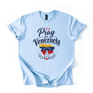 May include: A light blue short-sleeve t-shirt featuring the dark blue text Pray for Venezuela above a graphic of the Venezuelan flag in the shape of the country, with a pair of praying hands and a small red heart icon centered below.