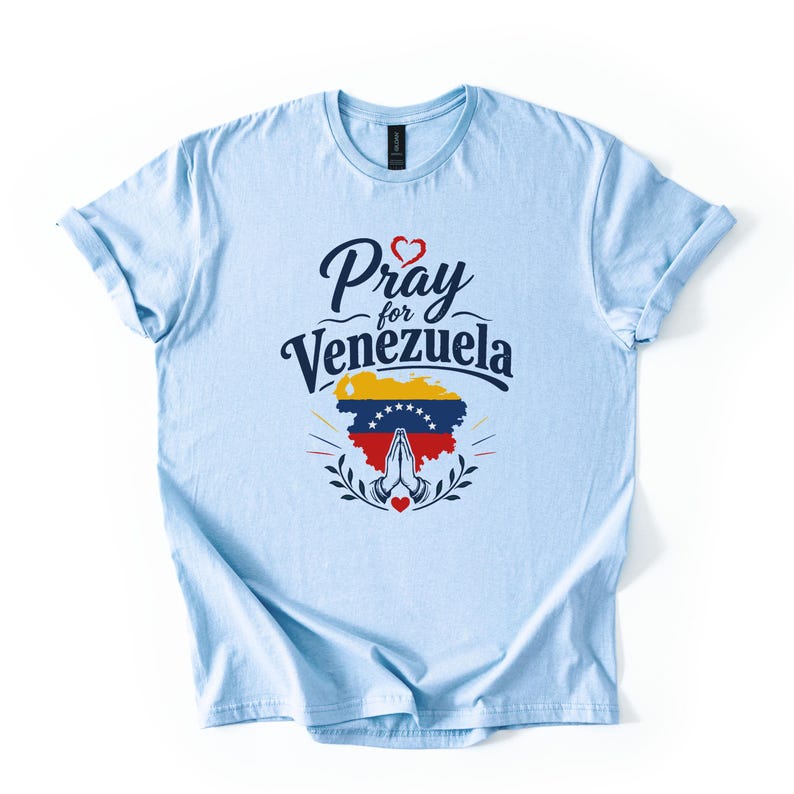 May include: A light blue short-sleeve t-shirt featuring the dark blue text Pray for Venezuela above a graphic of the Venezuelan flag in the shape of the country, with a pair of praying hands and a small red heart icon centered below.