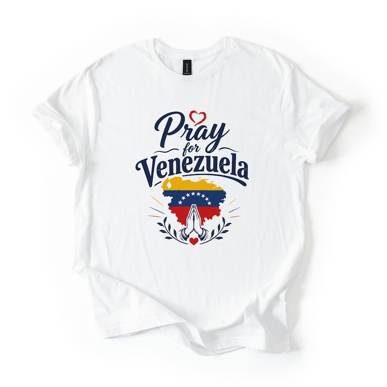 May include: A white short-sleeved t-shirt featuring a graphic print with the text Pray for Venezuela in dark blue. Below the text is a map of Venezuela filled with the national flag colors, yellow, blue, and red, with white stars and praying hands.